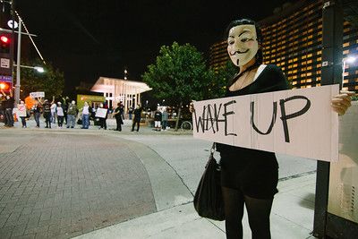 protestor at Anonymous protest wearing Anonymous mask and holding a sign that says "wake up."